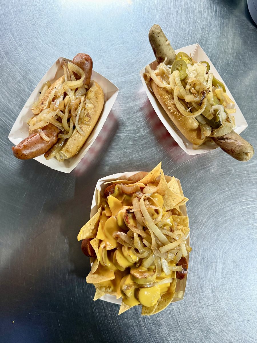 Two sausages on rolls with grilled onions, one also with jalapeños and sauerkraut, beside a boat of nachos with cheese sauce, sausage and grilled onions